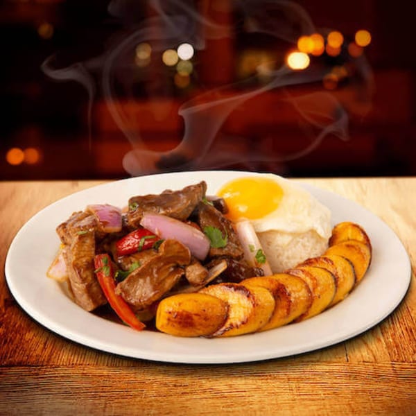 Steaming Peruvian Lomo Saltado with rice, fried egg, and plantains on a white plate.