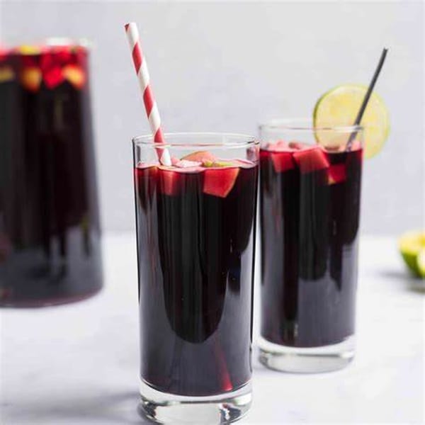 Two glasses of dark purple chicha morada with fruit, one with a striped straw, the other with a lime.