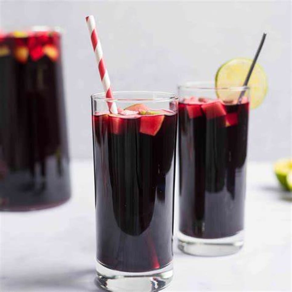 Two glasses of dark purple chicha morada with fruit, one with a striped straw, the other with a lime.